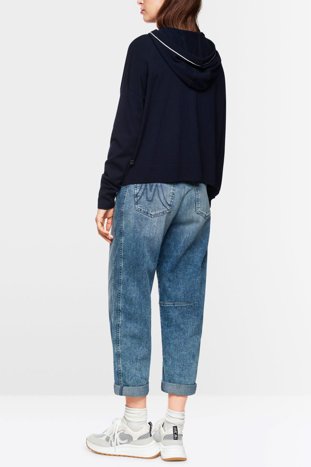 Femme portant un cardigan tricoté à capuchon zippé, associé à un jean décontracté et des baskets modernes.
