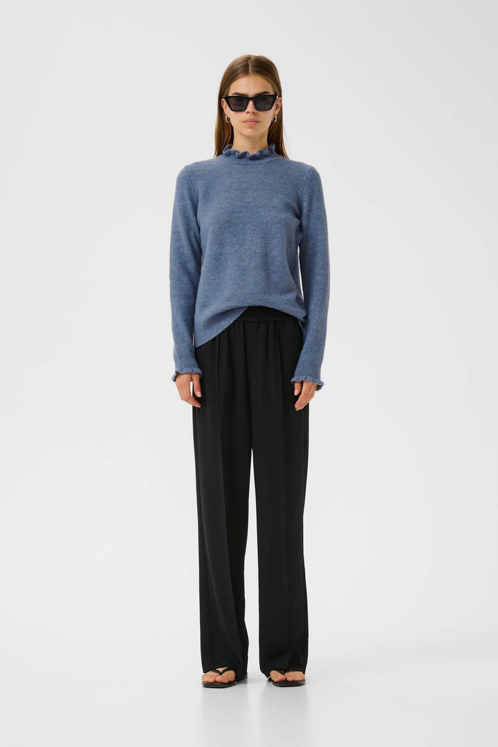 Femme portant un tricot Guni bleu à col montant froncé avec pantalon fluide, look chic et décontracté.
