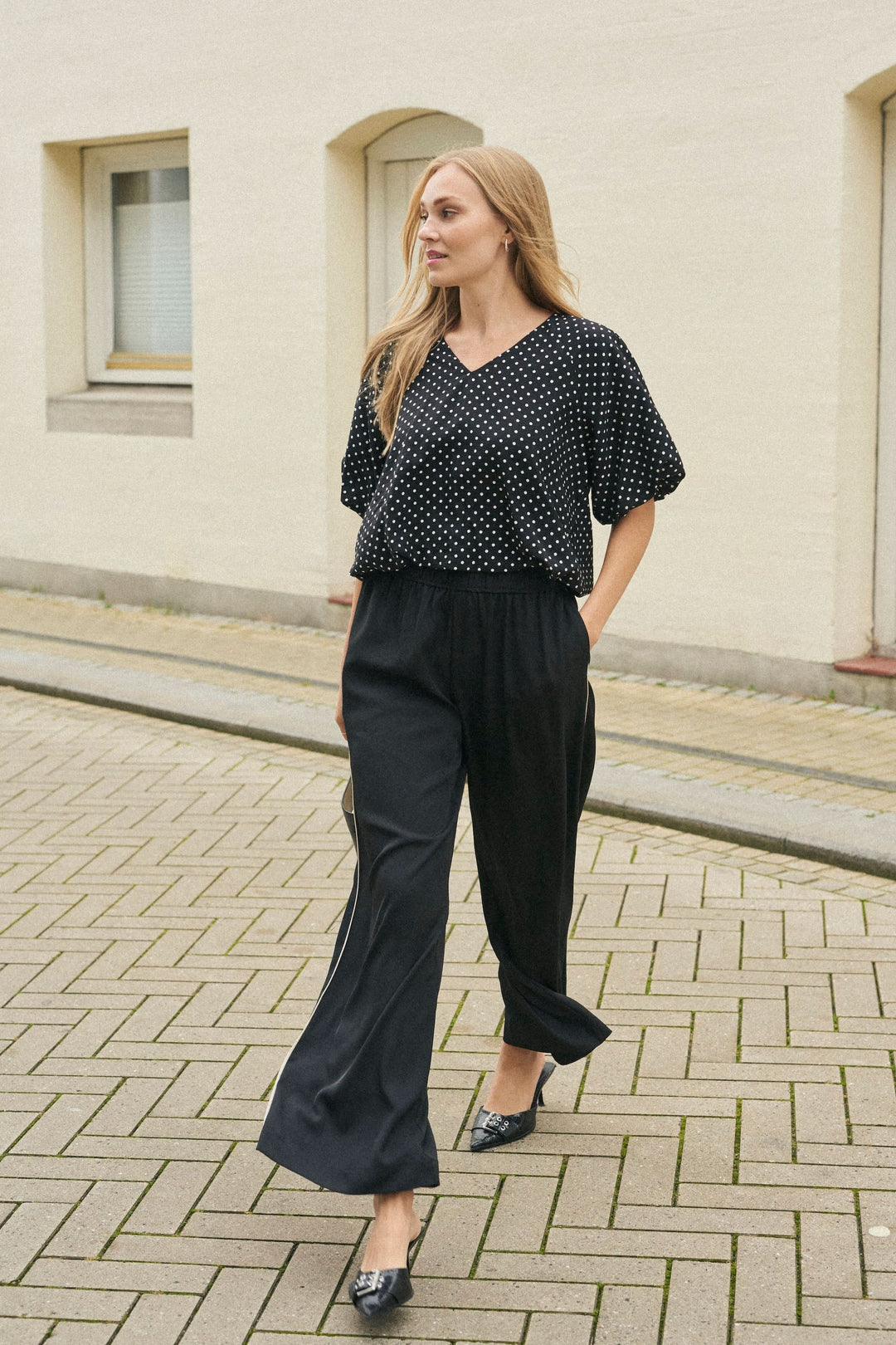 Femme marchant dans un pantalon fluide noir avec une taille élastique et un haut à pois, sur un chemin pavé.