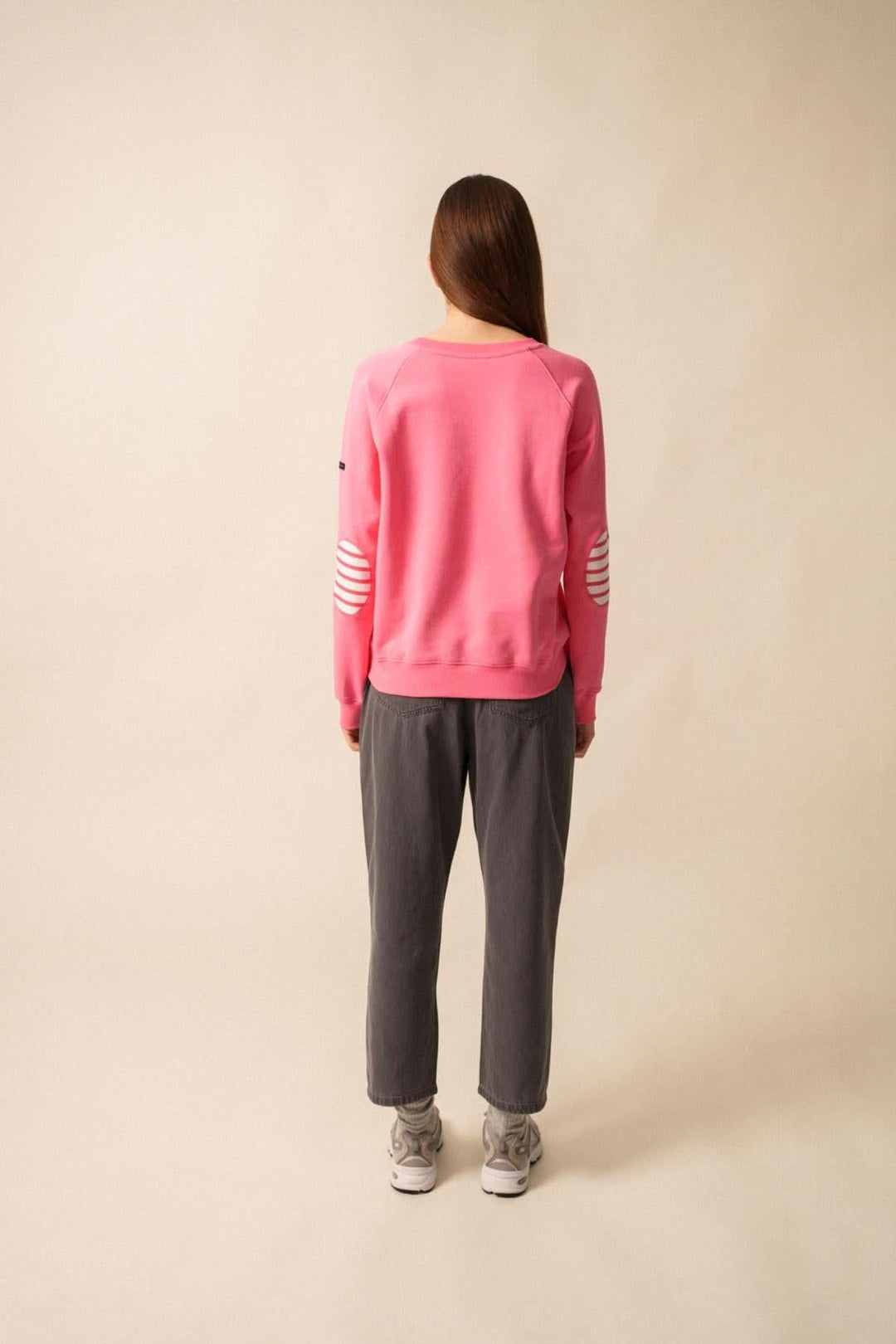 Femme de dos portant un sweat rose avec des coudières rayées et un pantalon gris, style casual.
