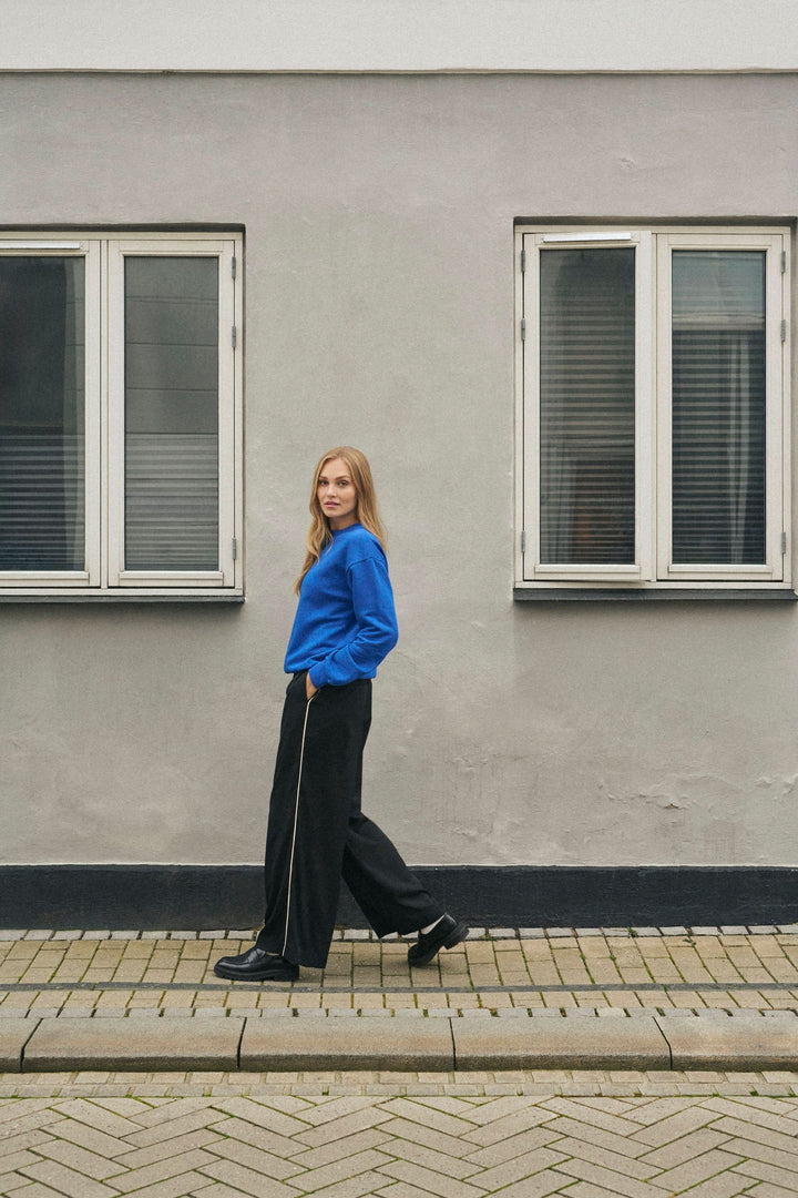 Femme en pantalon fluide noir et tee-shirt bleu posant devant un mur avec fenêtres.