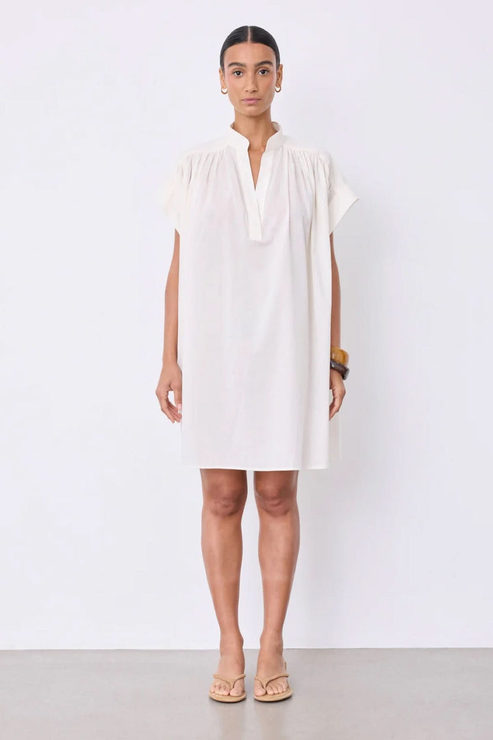 Robe Fani courte fluide col ouvert en blanc avec manches courtes et col en V, idéale pour un style minimaliste, portée par une femme debout.