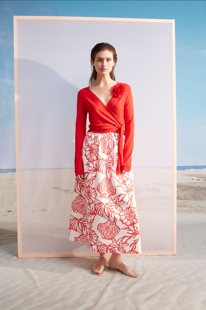 Modèle féminin portant un chandail portefeuille rouge avec décolleté en V et une jupe fleurie, sur fond de plage.