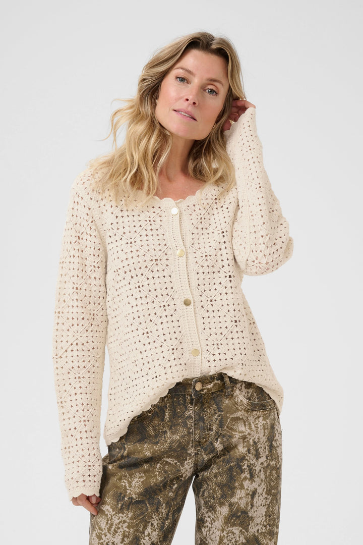 Cardigan ajouré beige avec boutons contrastants, porté avec pantalon à motifs, style rétro et contemporain.