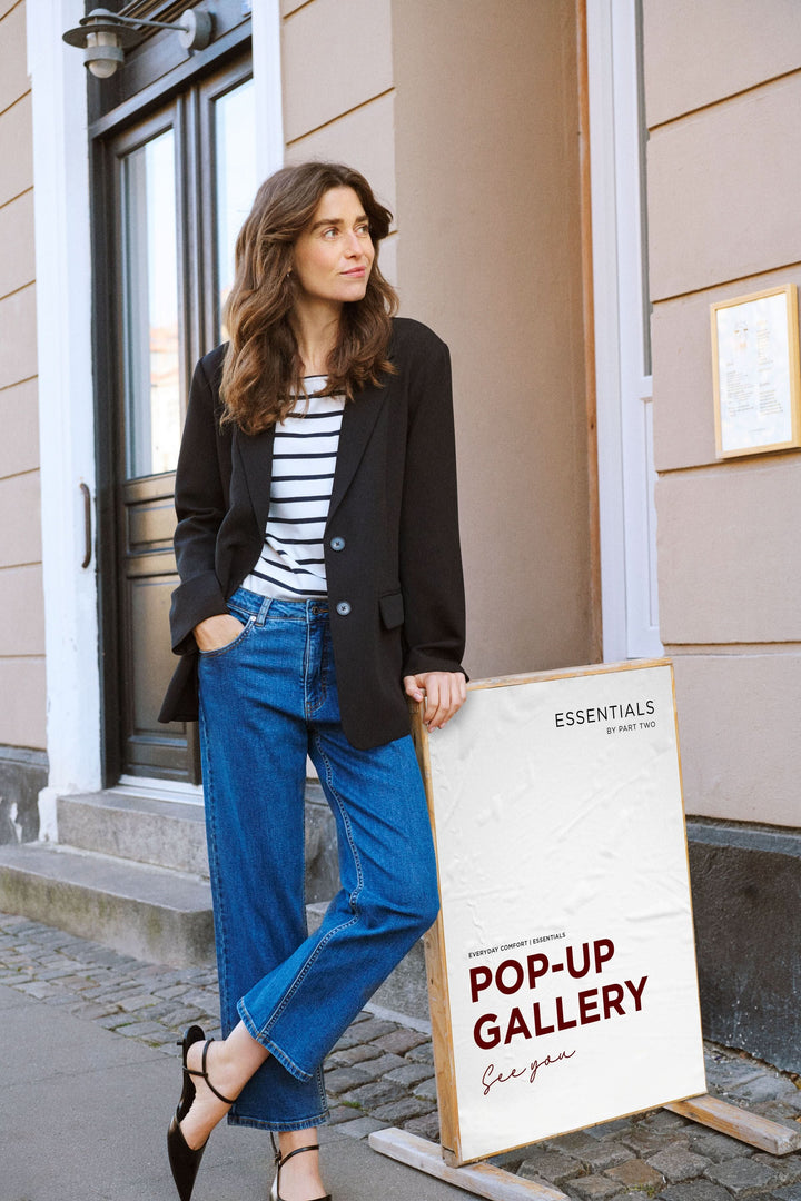 Femme en blazer noir et jean décontracté devant une galerie, style moderne et chic.