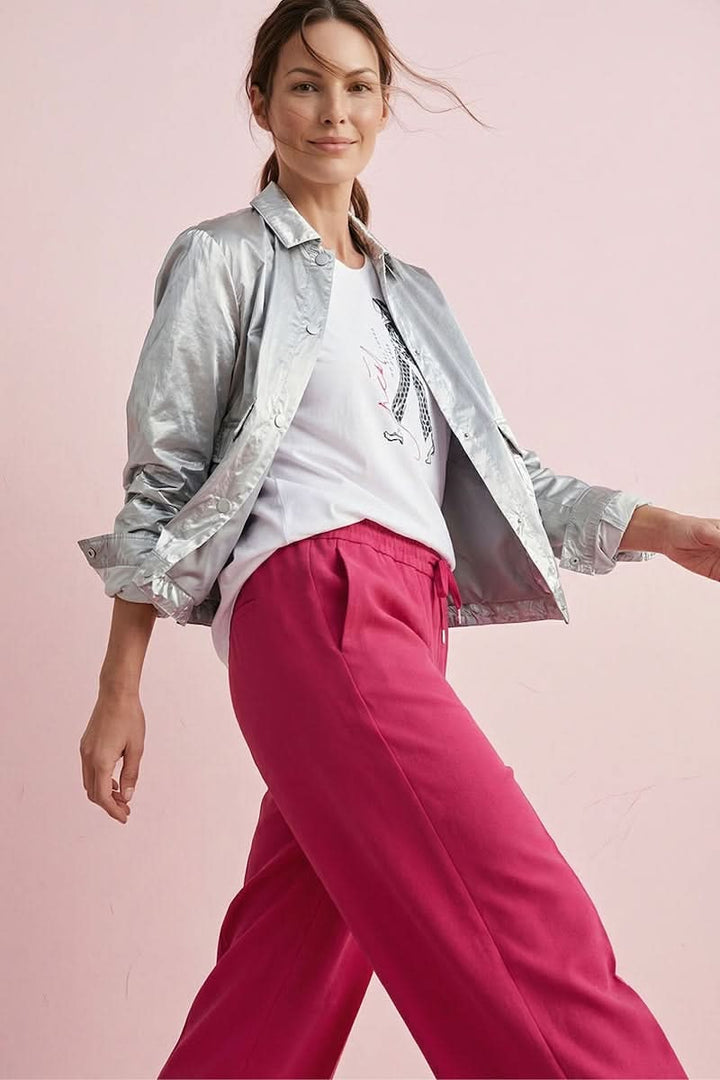 Femme portant un pantalon jogging fluide rose avec une veste argentée élégante devant un mur rose.