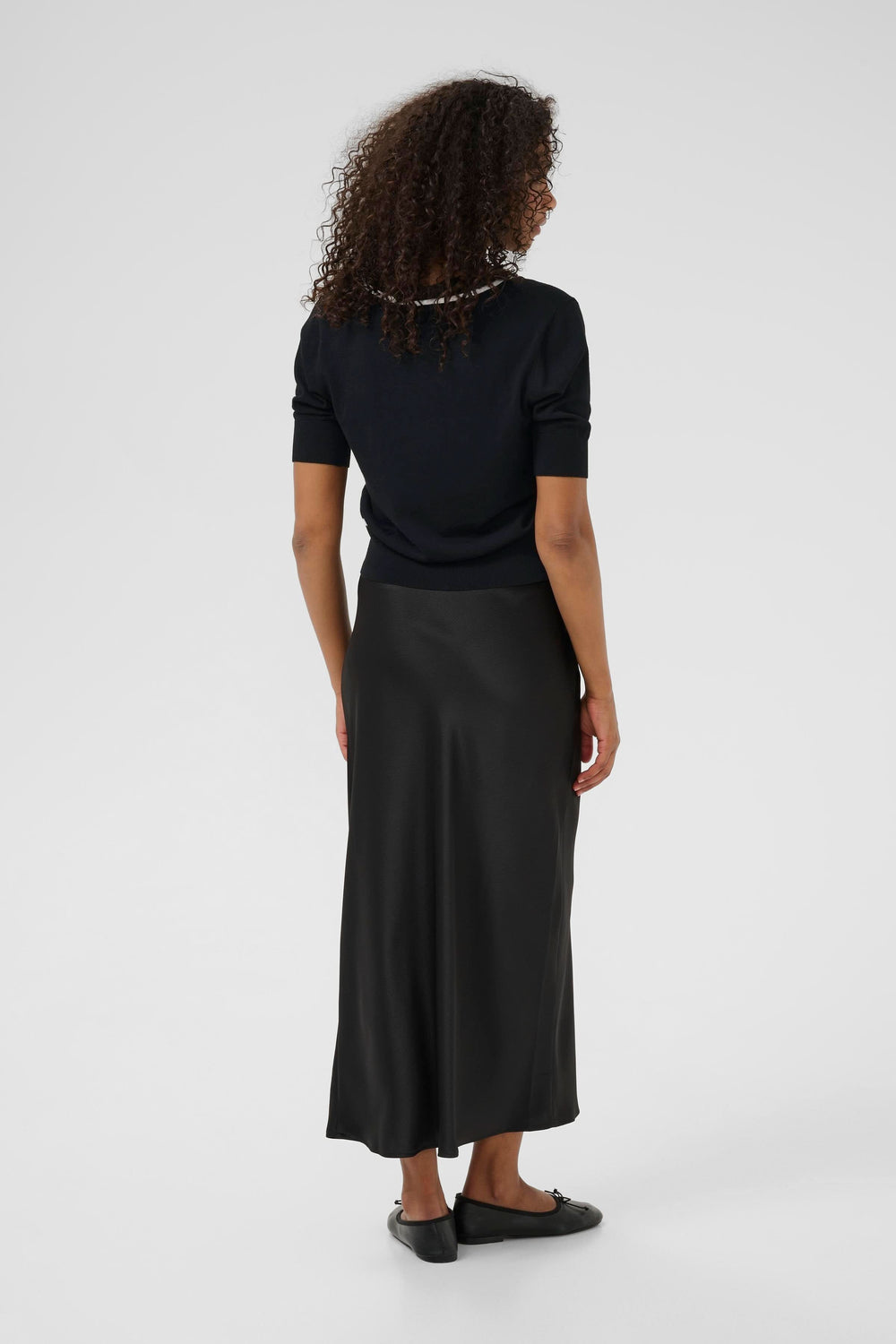 Femme de dos portant un cardigan court à manches courtes et une jupe longue noire, style chic et moderne.