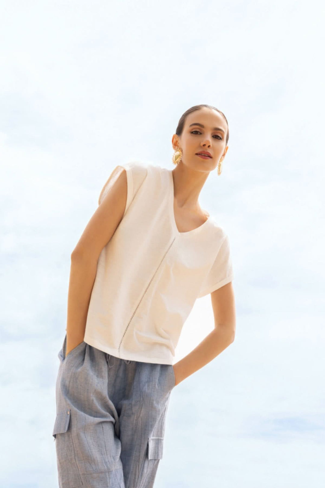 Femme portant un haut fluide col en V blanc avec manches courtes, posé de manière décontractée contre un fond ciel bleu lumineux.