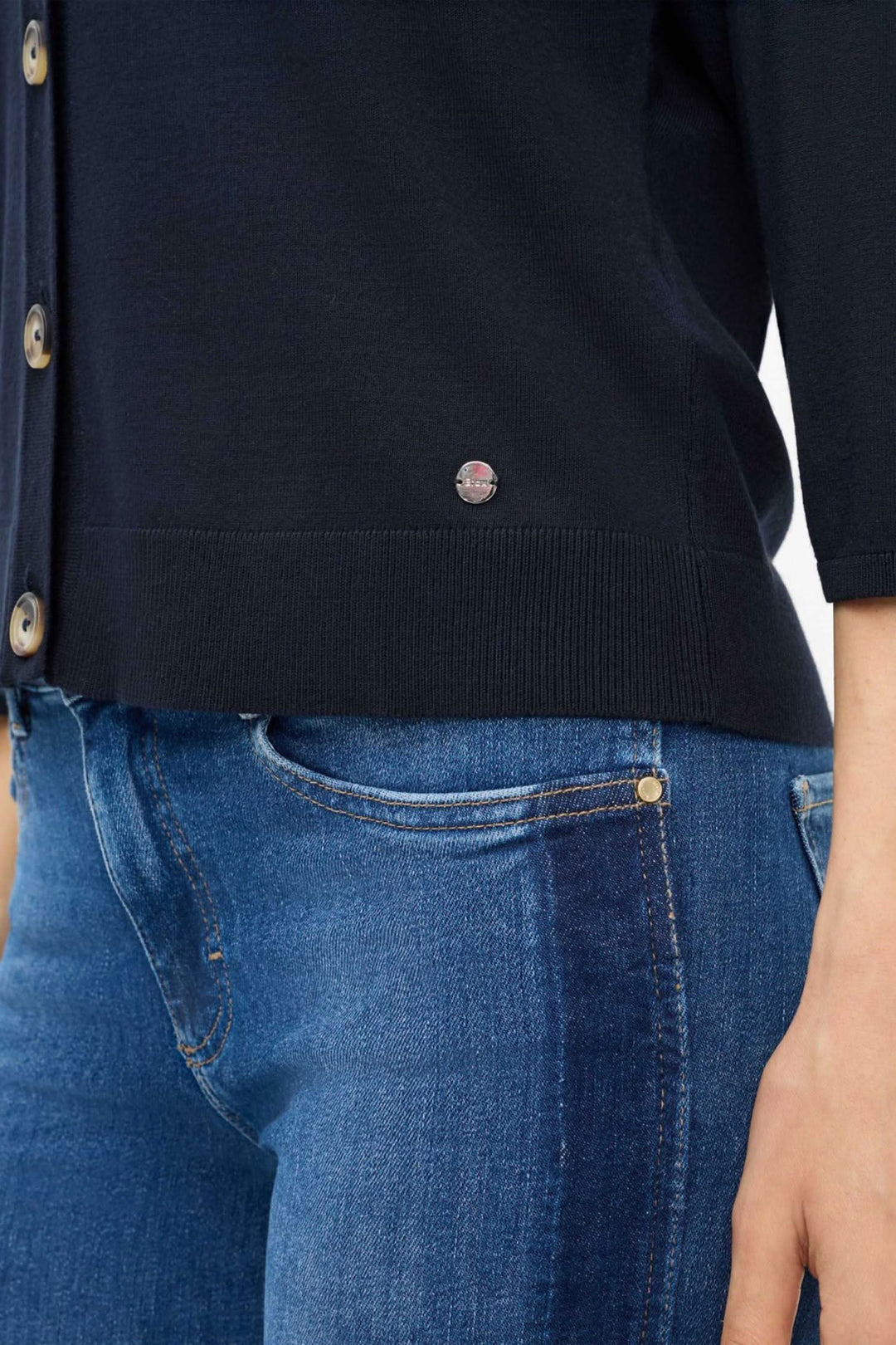 Détails du cardigan Anique avec boutons et logo, porté avec un jean bleu, mettant en avant la coupe moderne.