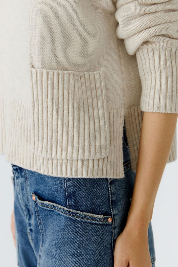 Chandail beige en maille fine avec poche plaquée et finitions côtelées, associé à un jean bleu.