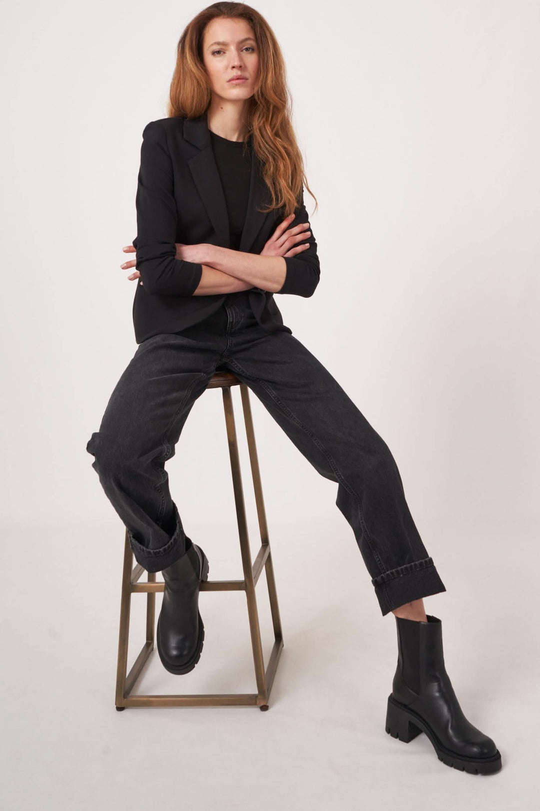 Femme assise sur une chaise, portant un veston en jersey stretch avec un haut noir et des bottines noires.