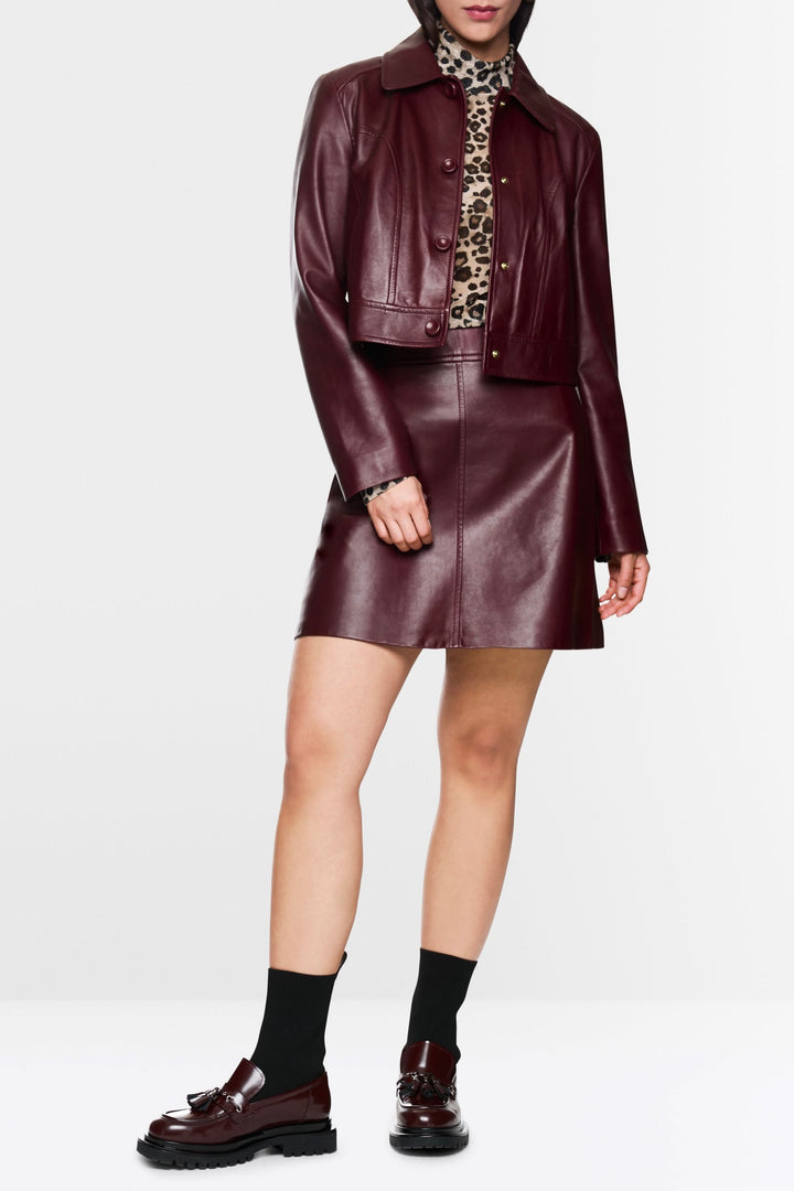 Veste en cuir bordeaux courte avec un look moderne, portée avec une jupe assortie et un col roulé imprimé.