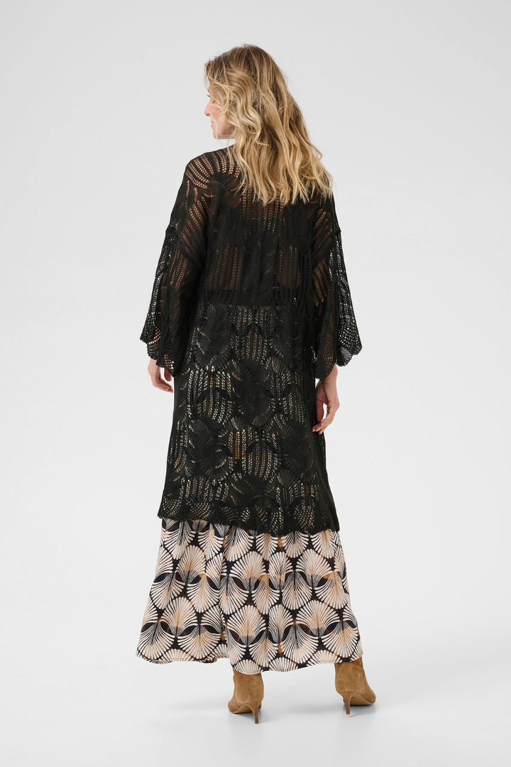 Cardigan Franca long en tricot ajouré noir, fluide, porté sur une jupe à motifs floraux.