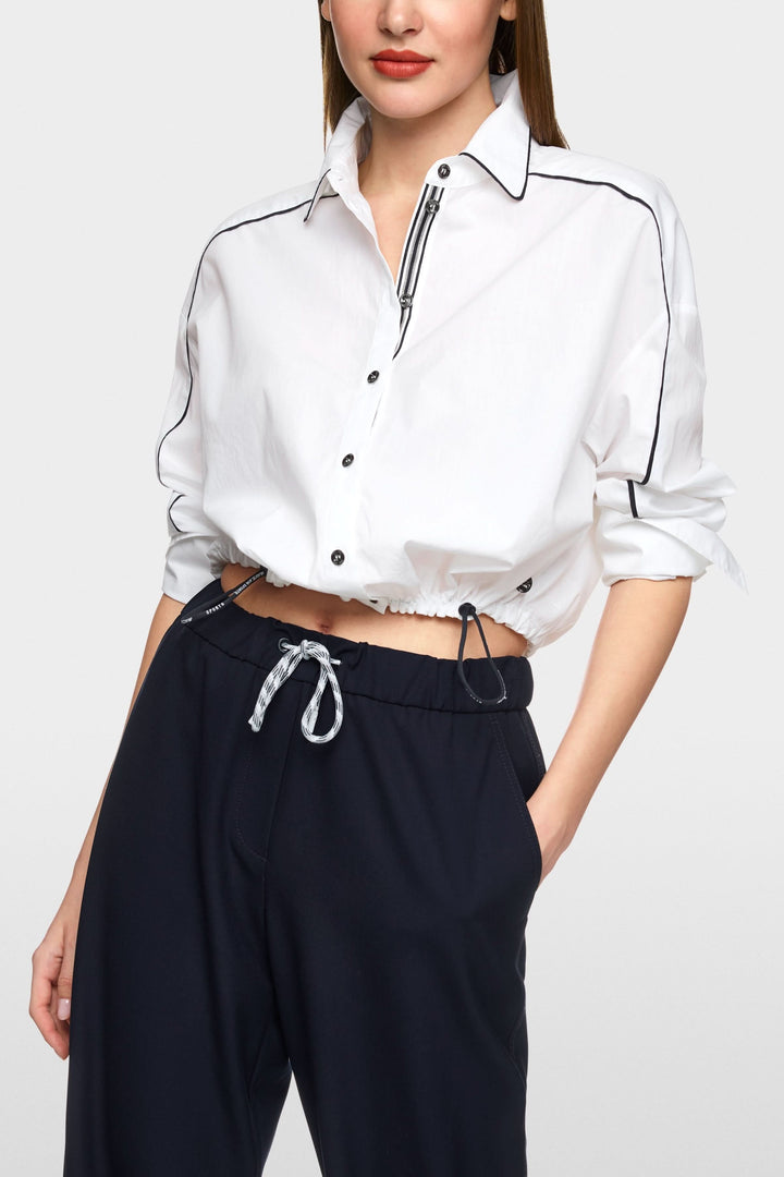 Chemise courte blanche avec détails contrastants, coupe ample et cordon ajustable à la taille, portée sur un pantalon noir.