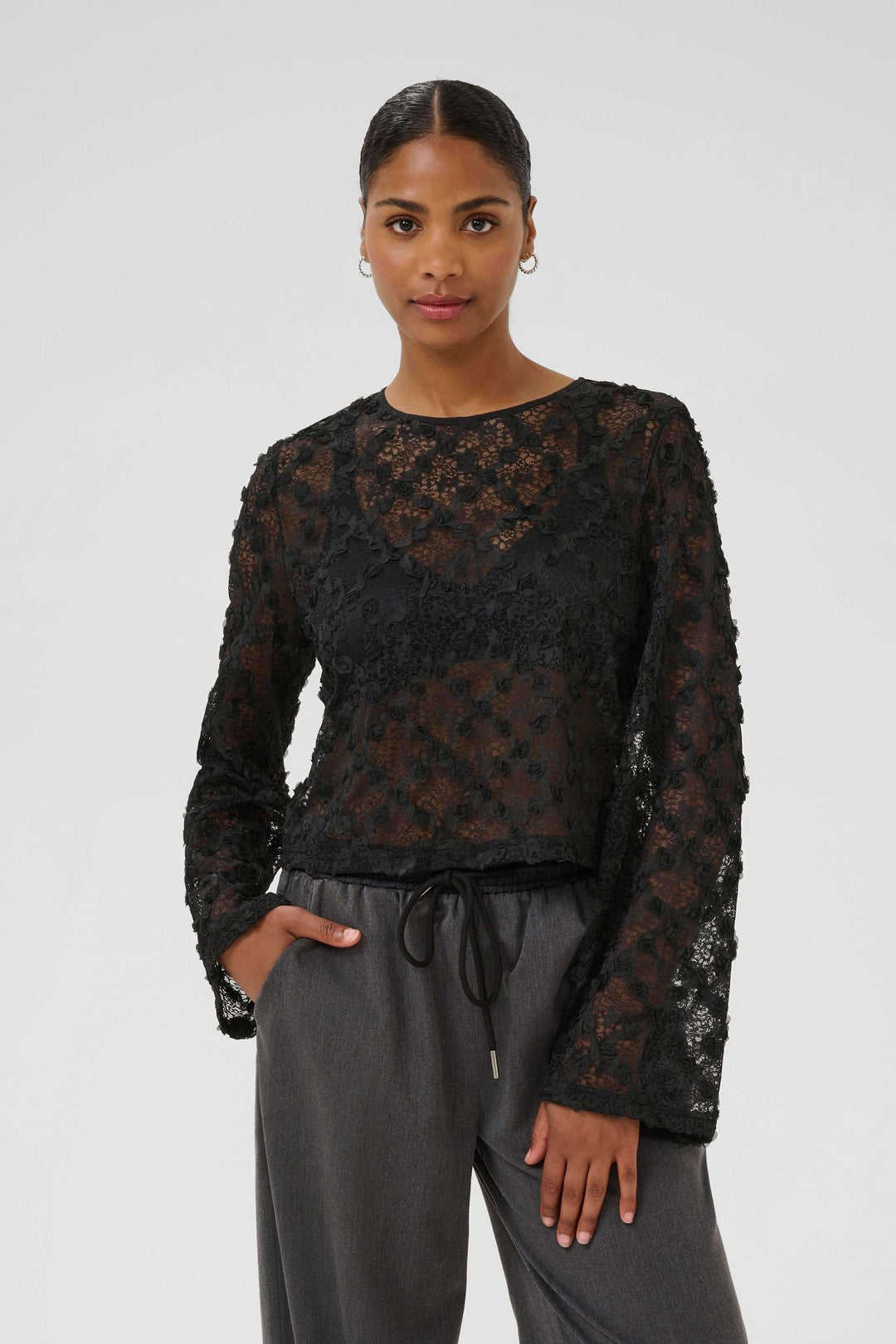 Femme portant la Blouse Rosie en dentelle florale à manches longues, élégante et moderne, avec dentelle transparente et manches évasées.