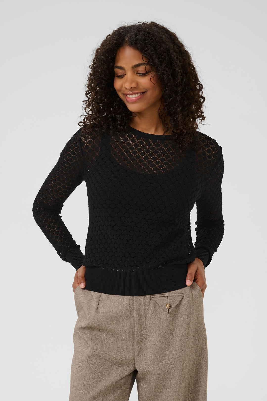 Chandail en maille ajourée noire avec motif géométrique, manches longues et encolure ronde, porté avec un pantalon beige.