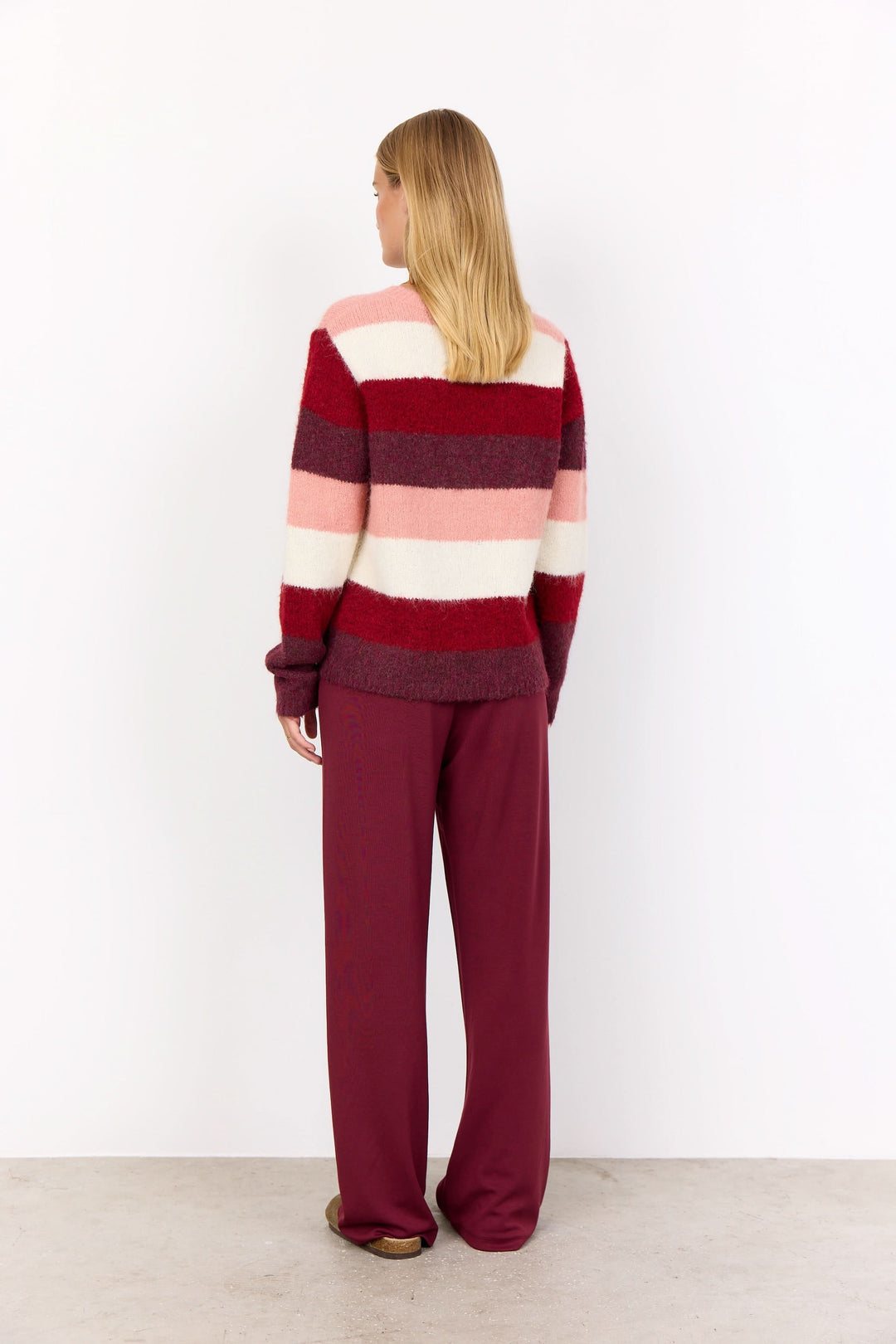 Femme de dos portant un tricot à rayures larges rouges et roses, associé à un pantalon unifié bordeaux.