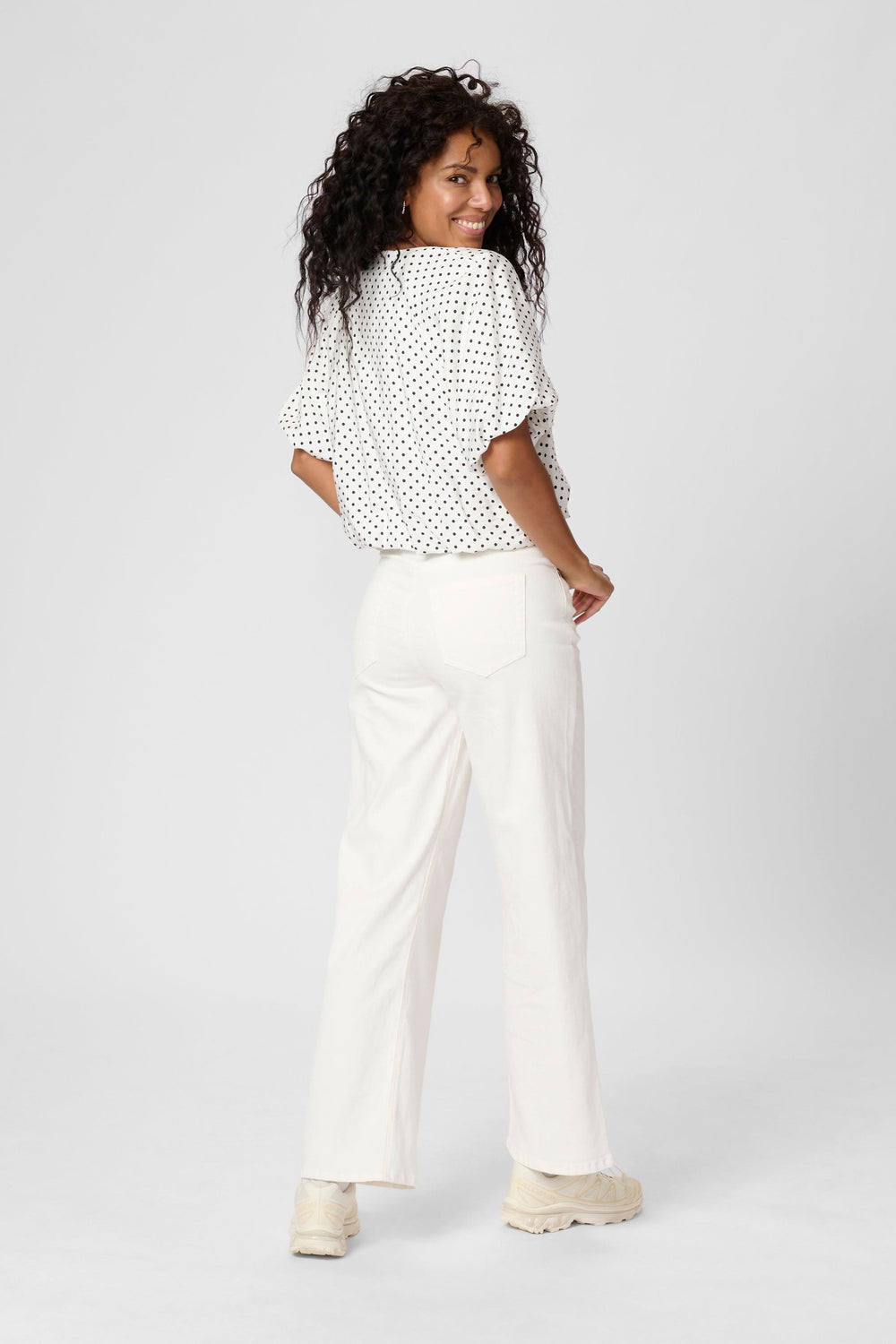 Femme portant un blouse à pois avec une encolure en V, vue de dos, associée à un pantalon clair.