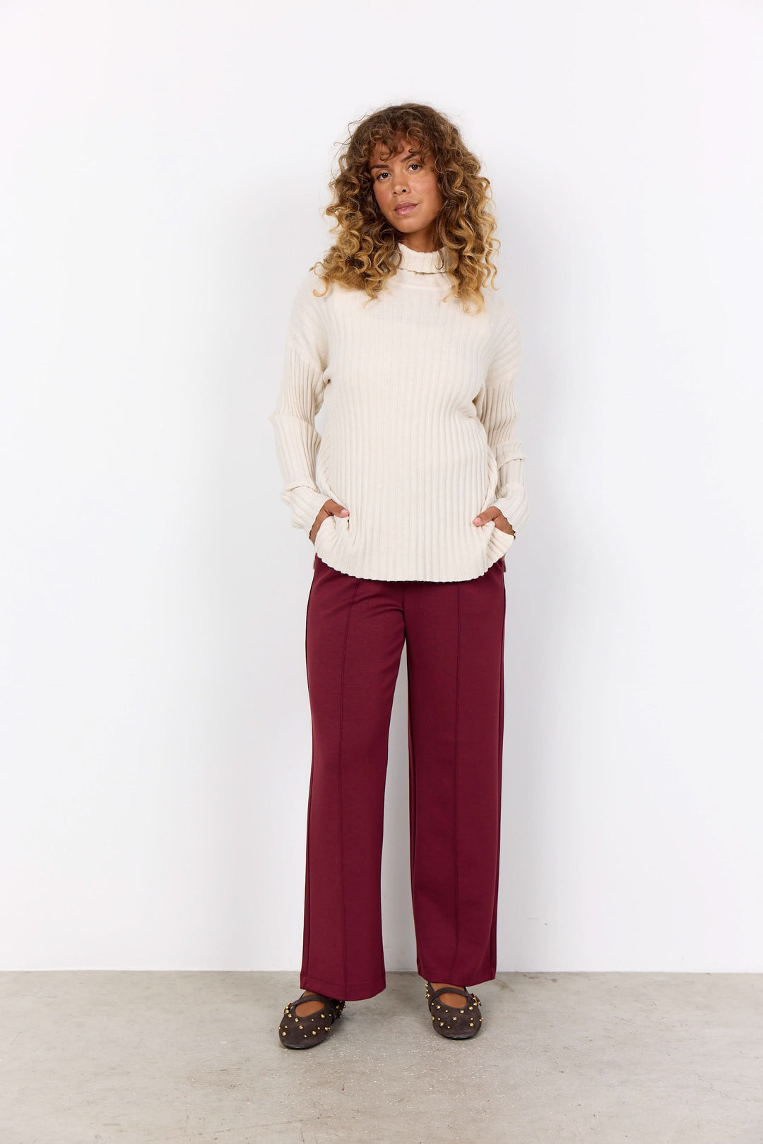 Femme portant un tricot col montant crème avec pantalon bordeaux, style décontracté et moderne.