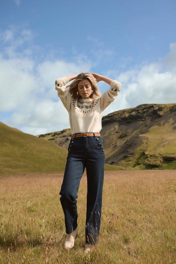 Femme en pull avec broderie, portant des jeans ajustés, posant dans un champ de collines verdoyantes sous un ciel bleu.