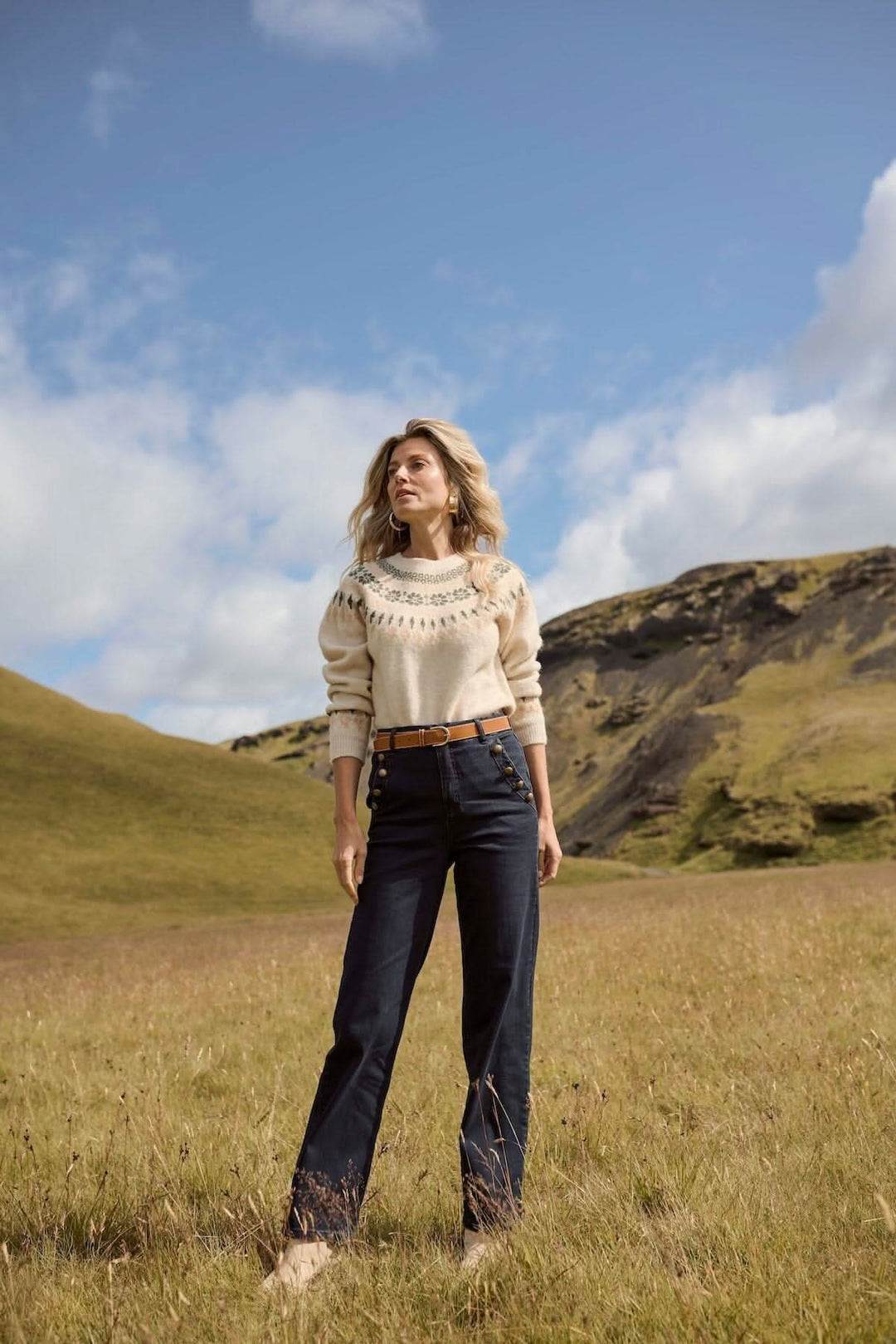 Femme portant un jean ajusté et un pull beige dans un champ à la campagne, avec un paysage montagneux en arrière-plan.